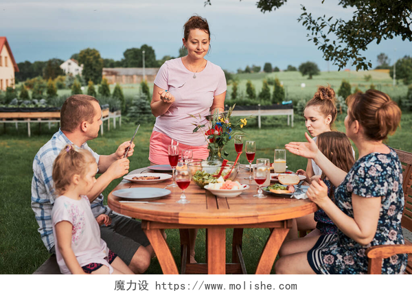 在一起聚餐的一家人夏天外出野餐时,一家人在自家花园的烤架边吃饭.围坐在餐桌旁，端着食物和碗碟的人